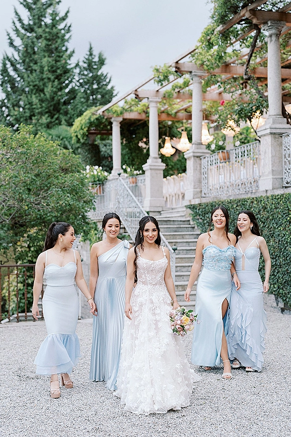 Bride with bridesmaids walking down a stone staircase, holding a pastel bouquet, under outdoor chandeliers by an ivy-covered pergola