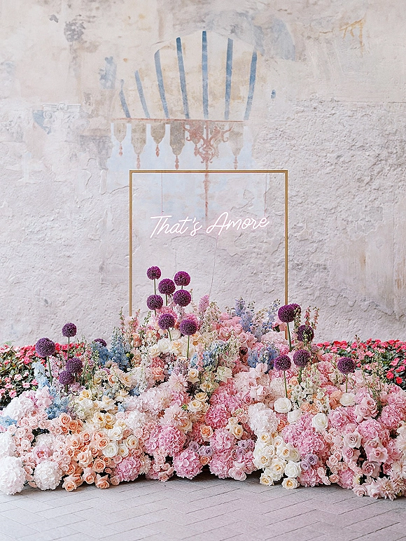 Wedding floral backdrop with neon wedding sign, pastel roses, hydrangeas and allium blooms over grounded florals on a stucco patio wall