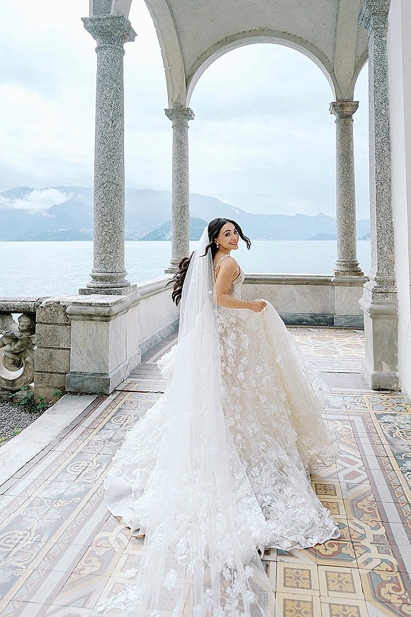 Bridal portrait of a bride looking back in a lace wedding dress with a long cathedral veil and train on a stone loggia by a mountain lake