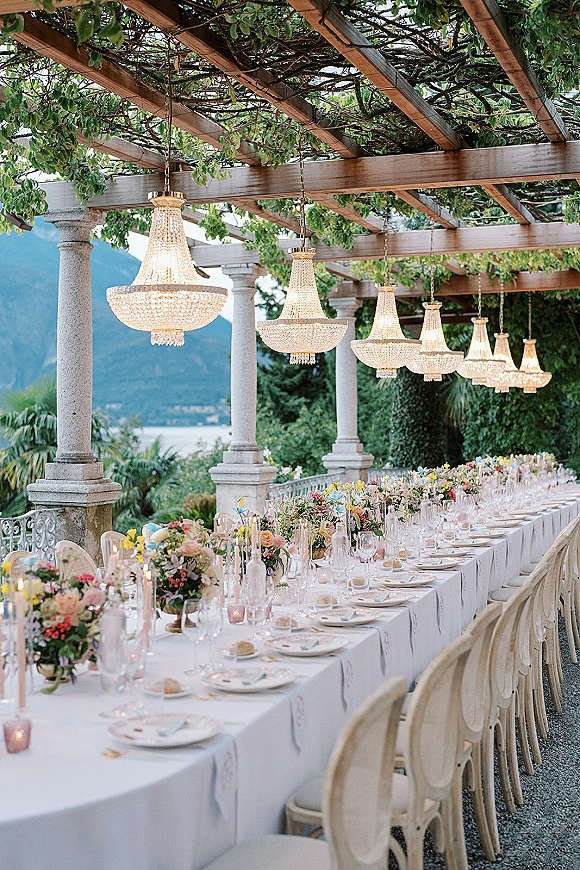 Reception tablescape with a long banquet table, white linens, floral centerpieces, and taper candles under crystal chandeliers on a lakeside terrace with mountains