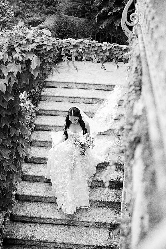 Bridal portrait of bride on staircase with a long veil train and bouquet, looking down on stone steps by an ivy wall and railing