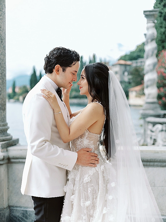 Wedding couple portrait of bride and groom embrace with a veil accent, forehead touch on a stone terrace overlooking a mountain lake and villa