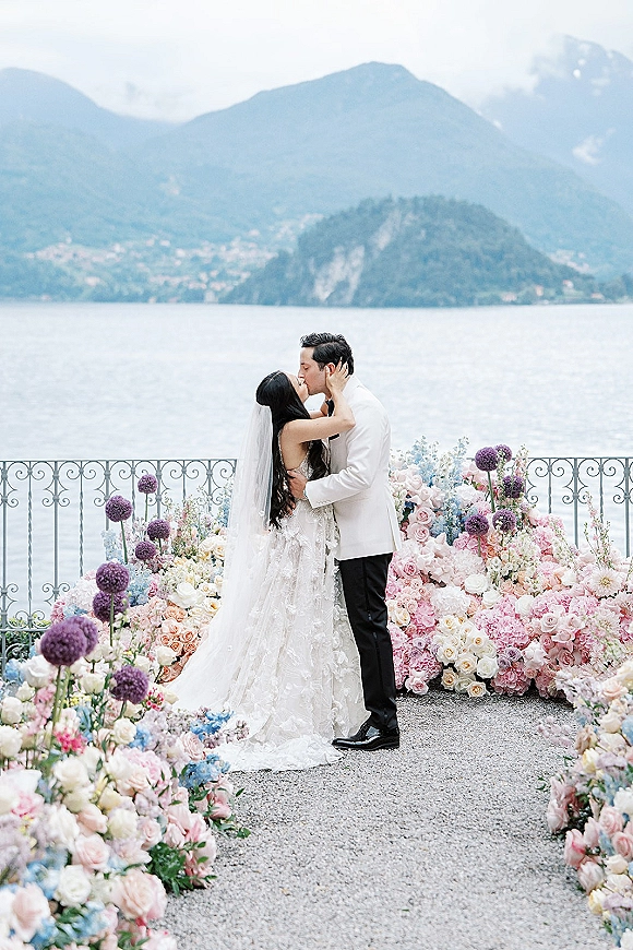 Wedding kiss as bride holds groom’s face, lace dress and veil flowing, on a stone terrace with lake and mountain backdrop