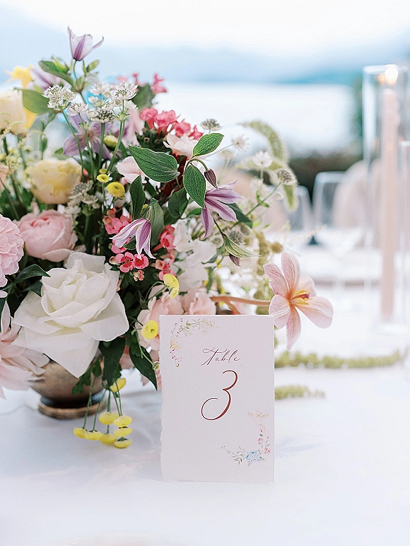 Reception tablescape with wedding table centerpiece, candles in glass holders and table number card on white linens by a waterfront view