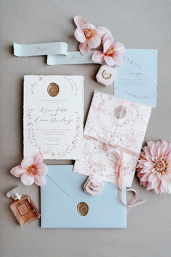 Invitation suite wedding invitation flat lay with deckle edge paper, vellum wrap, blush ribbon, wax seals, rings, and pink flowers on gray fabric
