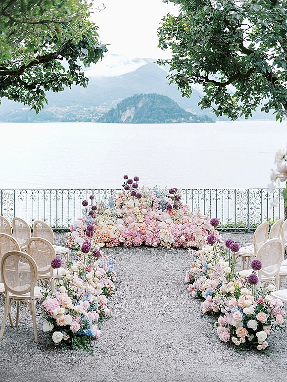Ceremony setup for an outdoor wedding ceremony with pastel aisle flowers and light wood chairs on a gravel terrace overlooking a lake and mountains