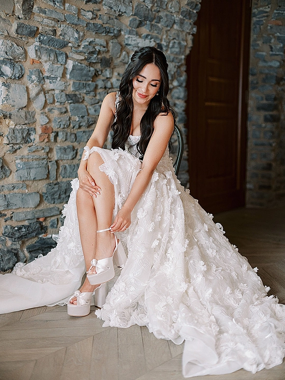 Bridal portrait of a bride putting on satin platform heels, seated on a wood floor in a lace and tulle gown by a stone wall