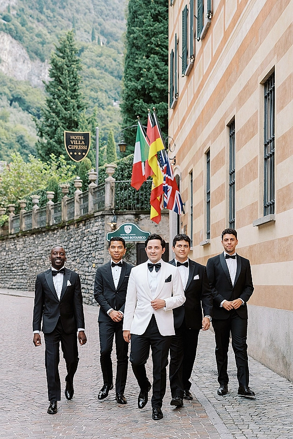 Groomsmen portrait with groom in white tux leading black-tie friends in bow ties, walking past a hotel stone wall on cobblestones