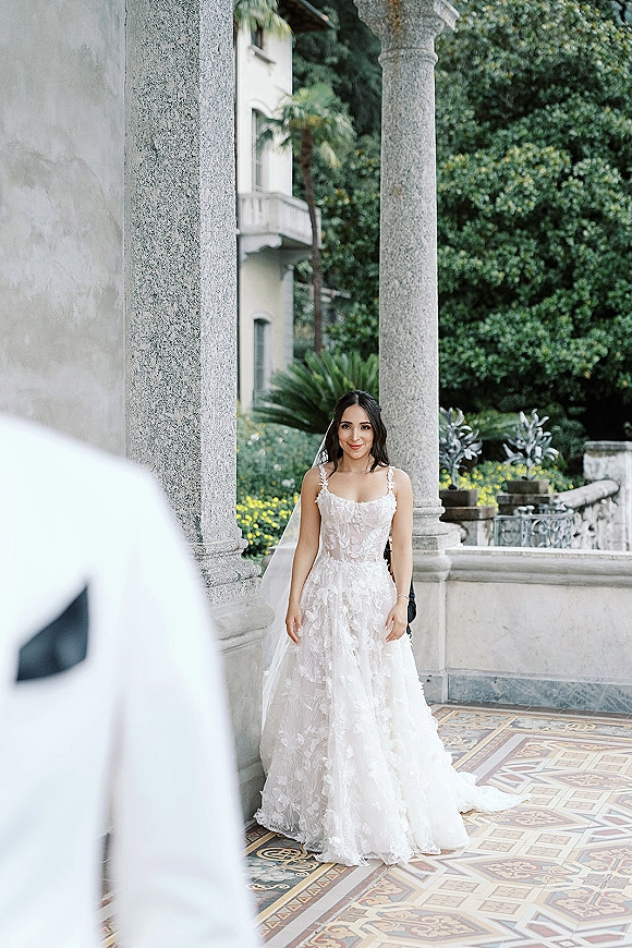 First look moment as the bride in a lace gown and veil approaches the groom on a villa terrace with stone columns and palms in back