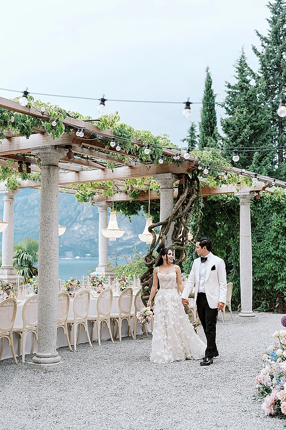 Couple portrait of bride and groom walking hand in hand under a vine-covered pergola with string lights by a lakeside mountain view