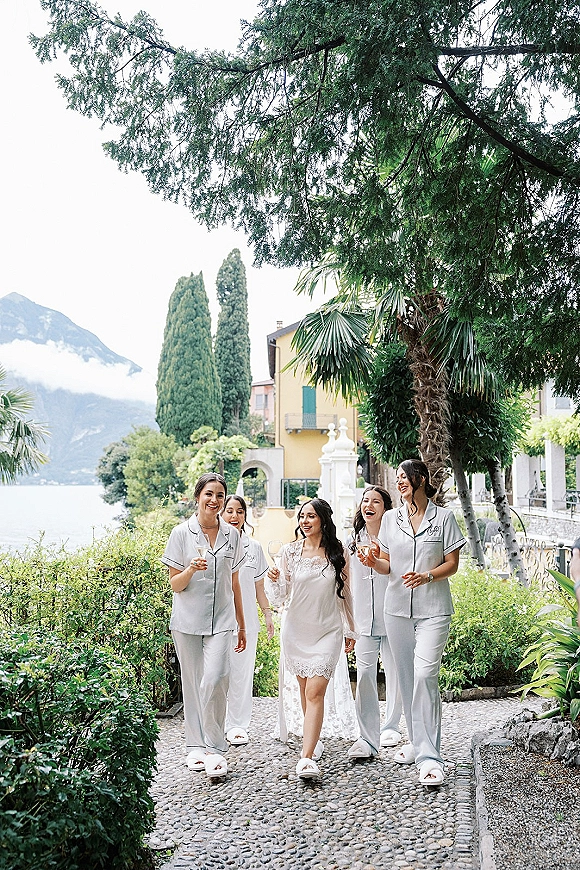 Bridesmaids getting ready in bridal party pajamas walk together holding champagne flutes along a villa garden pathway under overcast sky