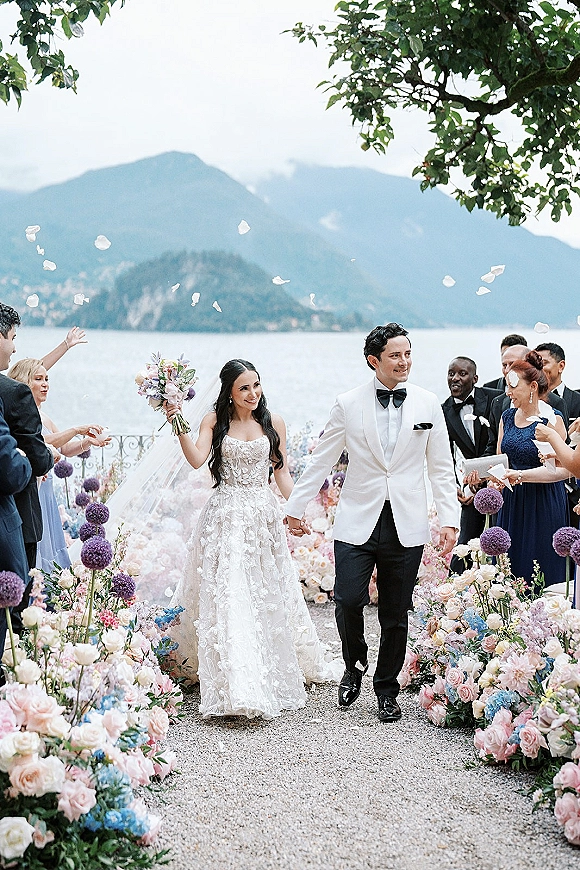 Wedding recessional as bride and groom walk the aisle holding hands, bouquet and veil flowing, guests toss rose petals by a mountain lake terrace