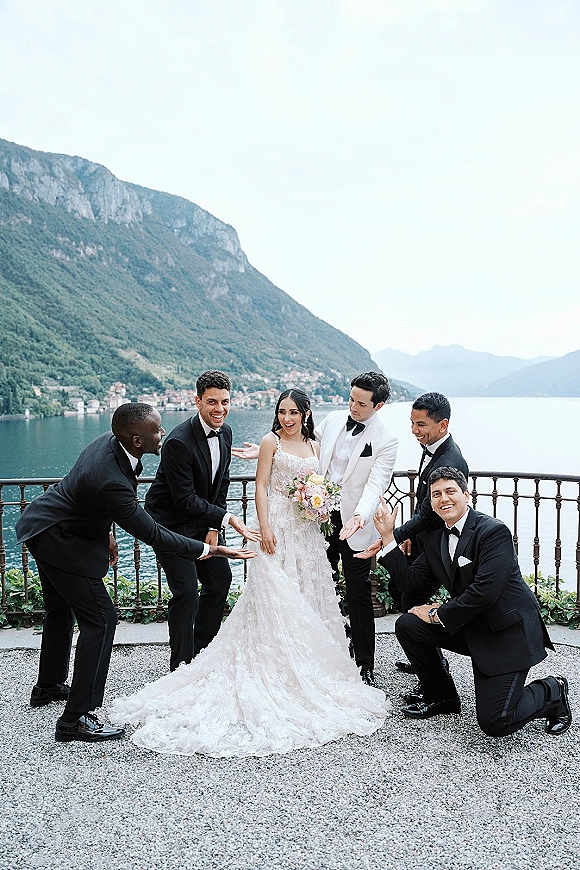 Wedding party portrait of bride with groomsmen in tuxedos, holding a pastel bouquet on a terrace with lake and mountains under cloudy sky