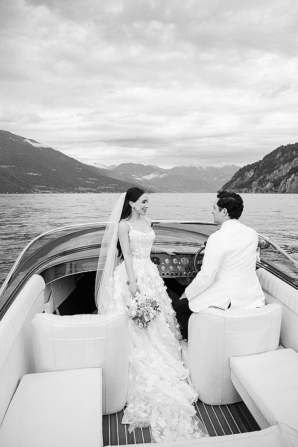 Couple portrait of newlyweds on boat, bride in veil holding bouquet beside groom, with mountain lake and cloudy sky behind