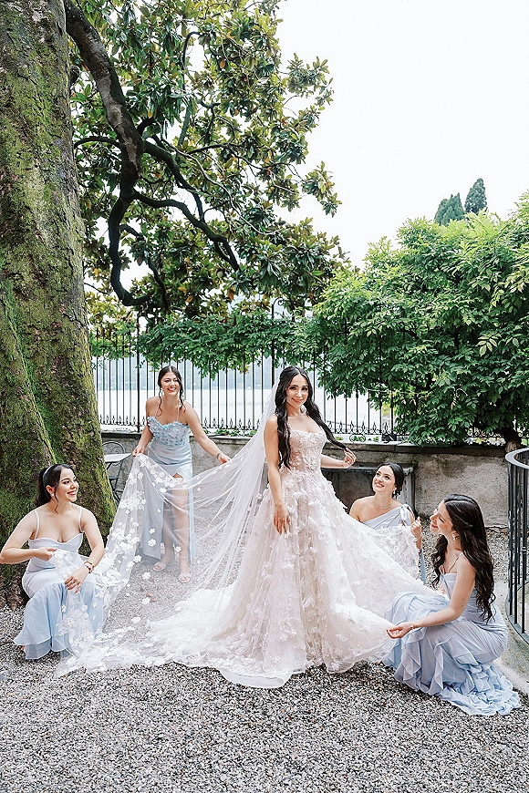 Bride with bridesmaids photo as attendants kneel to arrange her long lace veil in a garden courtyard beneath a large tree