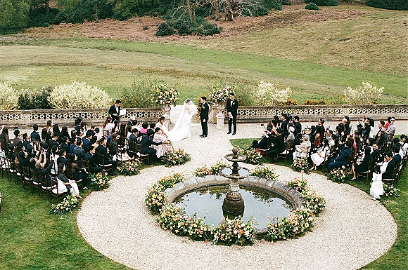 Outdoor wedding ceremony with bride and groom at a flowered arch, guests seated in a circular layout on a garden lawn by a fountain