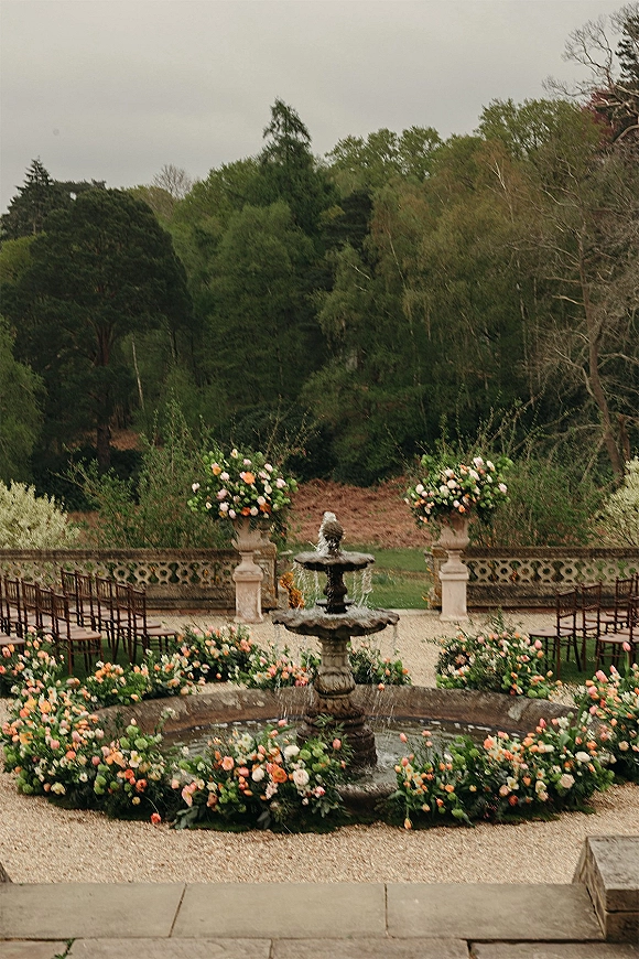 Outdoor ceremony setup with a garden wedding ceremony around a stone fountain, circular chairs and pedestal urn florals on a terrace