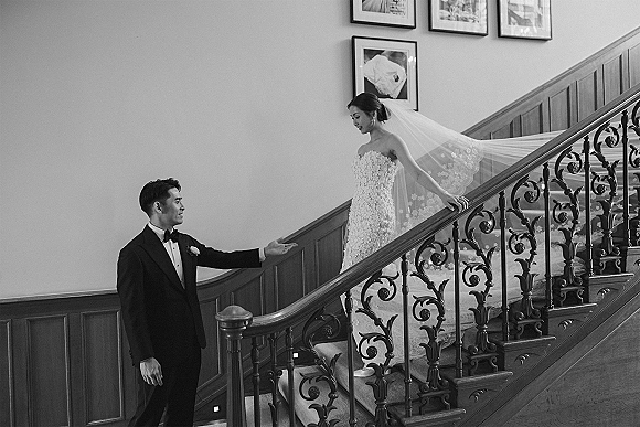 Wedding first look as groom reaches up to bride on staircase, her long lace veil trailing down carpeted stairs in wood-paneled interior