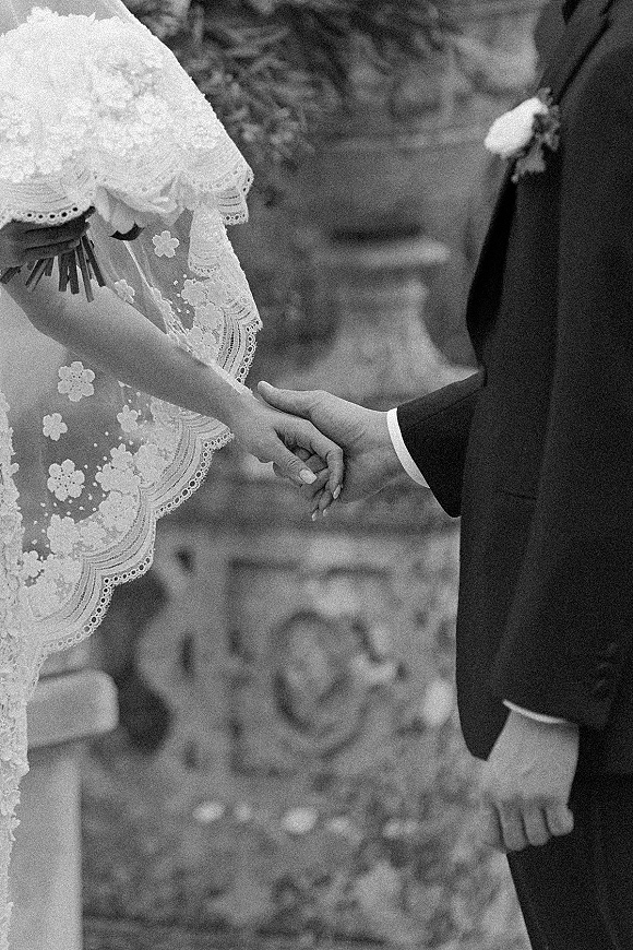 Ceremony handholding as bride and groom clasp hands, lace sleeve and veil framing a bouquet, with garden greenery and stone pedestal behind