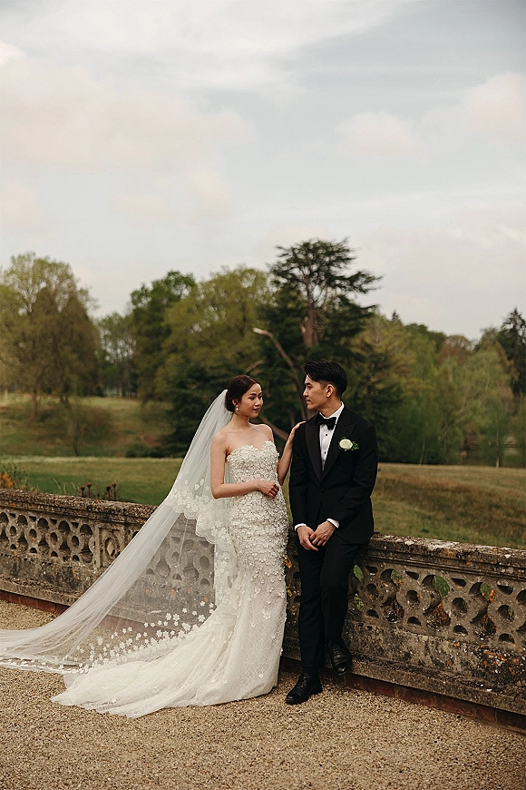 Couple portrait of bride and groom leaning on a stone balustrade, her long lace veil trailing over gravel path under cloudy sky
