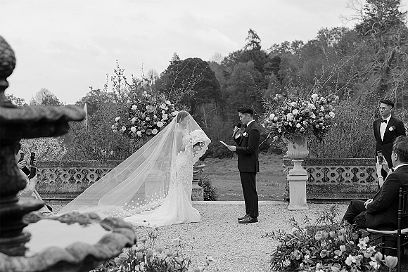 Wedding vows as groom reading vows into a microphone, bride in lace dress with bouquet and long veil on a garden terrace with floral urns