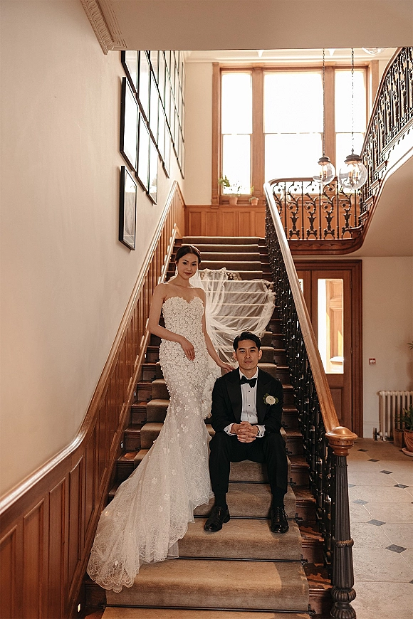 Couple portrait on a grand indoor staircase, bride in a strapless lace wedding dress with long veil, groom in tuxedo by wood paneling