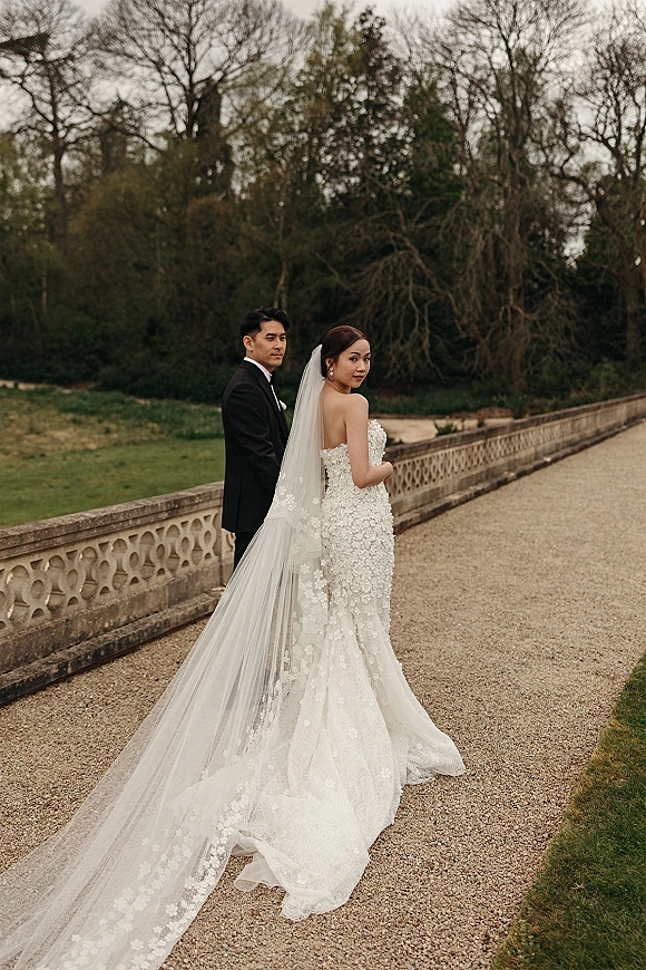 Couple portrait of bride in strapless gown looking back as groom in tux stands behind her, long veil train flowing by stone balustrade in garden