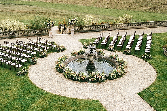 Outdoor ceremony setup with garden wedding ceremony seating in a circular layout, blush and peach florals lining a gravel aisle around a stone fountain.