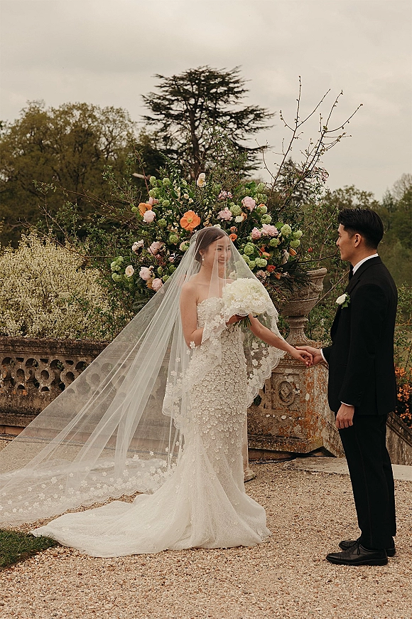 Couple portrait of bride and groom holding hands on a stone terrace, her cathedral veil and white bouquet beside an urn planter