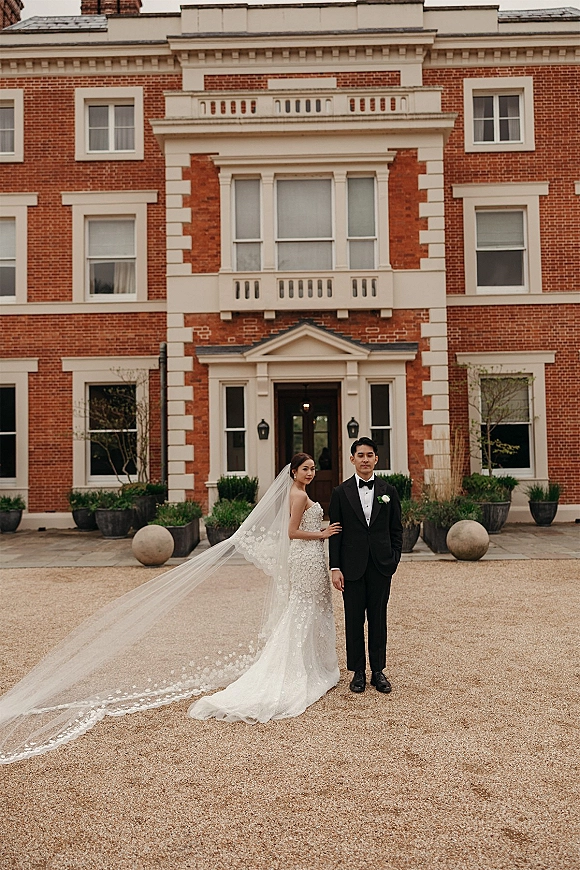 Couple portrait of bride and groom in black-tie attire, bride in lace dress with long veil train, posed by a brick courtyard entrance
