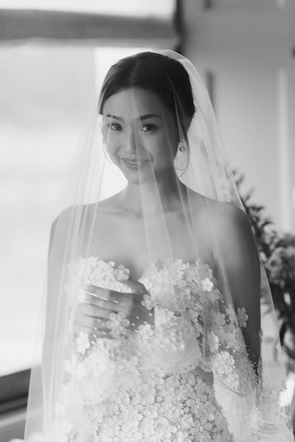 Bridal portrait of a bride in a strapless lace dress holding her veil over her face, lit by window light with blurred greenery behind