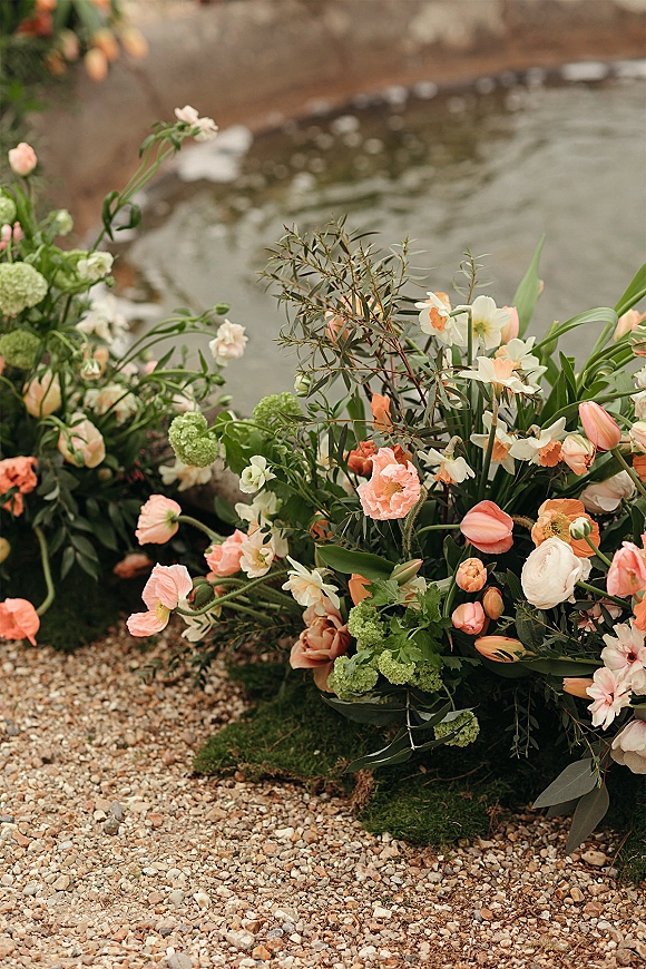 Wedding floral arrangement ground floral installation with tulips and ranunculus nestled in moss beside a stone fountain and gravel path