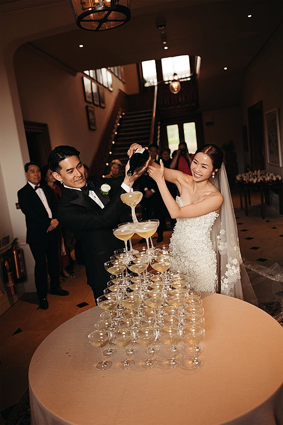 Champagne tower moment as bride and groom pour sparkling wine into stacked coupe glasses, guests watching in an indoor foyer by staircase