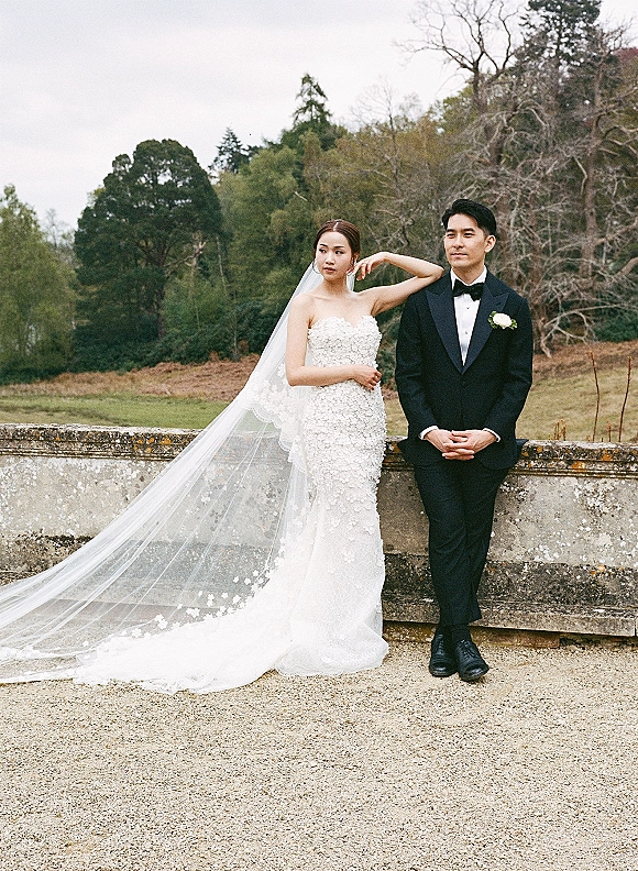 Couple portrait of bride in a strapless lace wedding dress and groom in tuxedo by a stone wall on an overcast garden lawn