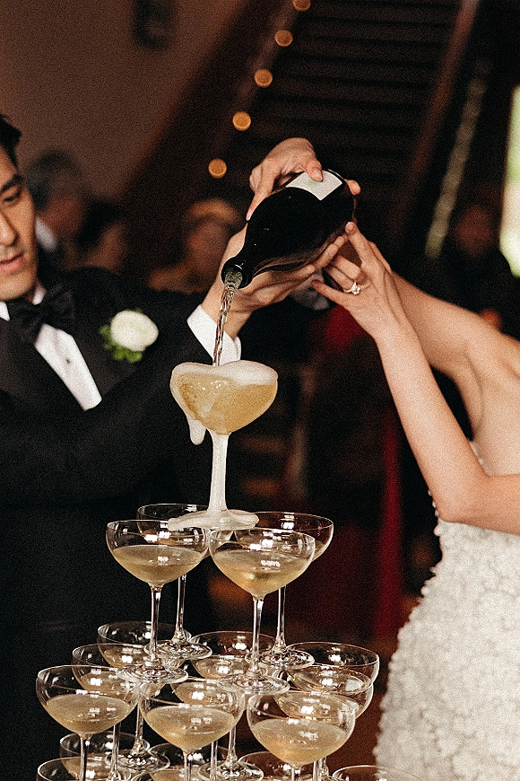Champagne tower pour as bride in bridal gown and groom in tuxedo pour bubbly into coupe glasses under string lights at reception