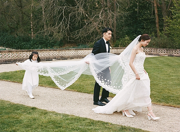 Wedding couple portrait with bride walking as her lace veil trails behind, groom in black tuxedo beside her on a garden path