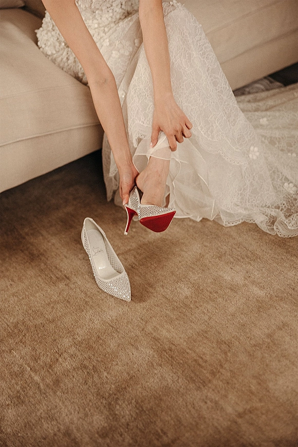 Bridal shoes with crystal high heels and a red sole beside a lace wedding dress on a beige sofa, getting-ready details on carpet floor