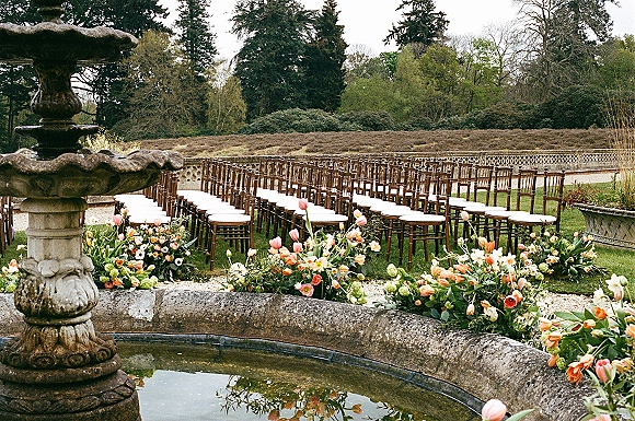 Outdoor ceremony setup with garden wedding ceremony aisle lined with orange and pink tulip florals, chiavari chairs, and a stone fountain backdrop