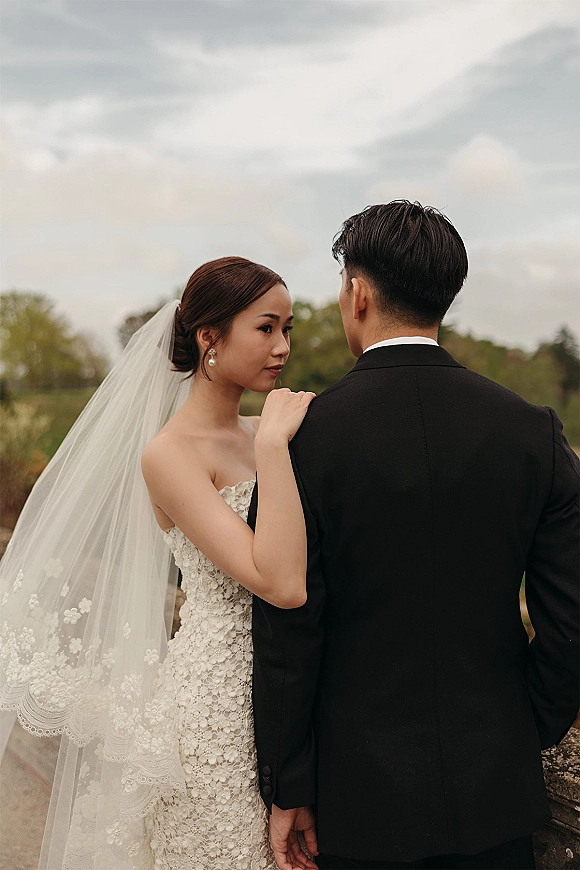 Couple portrait of bride and groom embrace, bride looking over shoulder in lace strapless gown and veil beside a stone ledge field