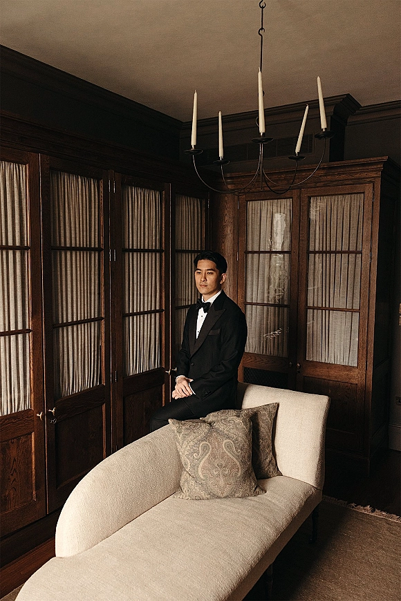 Groom portrait in a black tie tuxedo with bow tie and boutonniere, seated on a cream chaise in a wood-paneled room under a chandelier