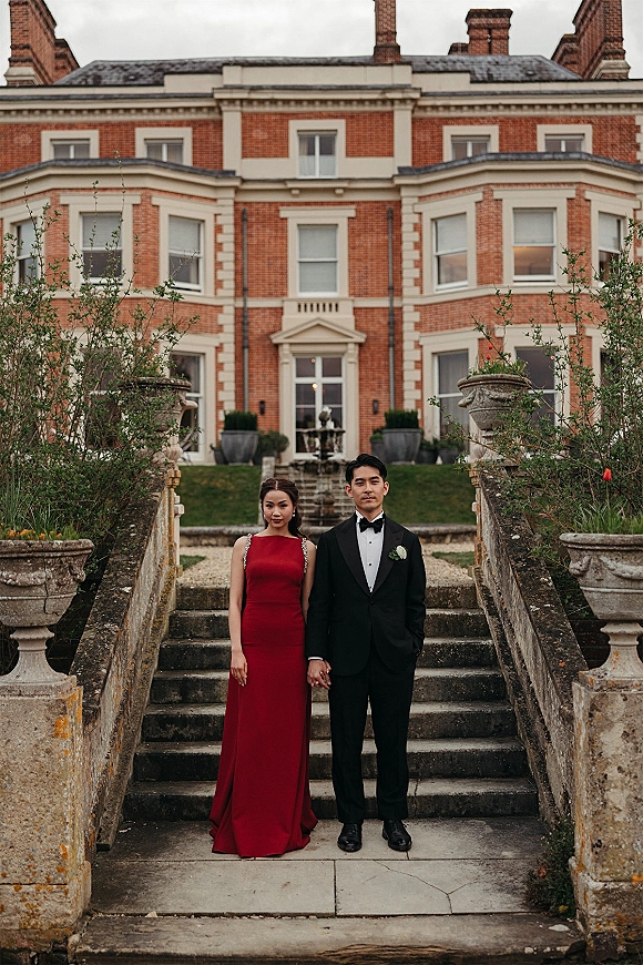 Couple portrait of bride in a red evening gown holding hands with groom in black tux on a stone staircase at a brick manor house