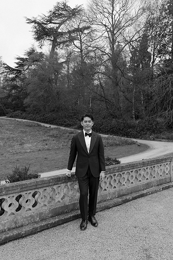 Groom portrait in a classic black tuxedo with bow tie, standing on a gravel garden path by a stone balustrade under overcast sky