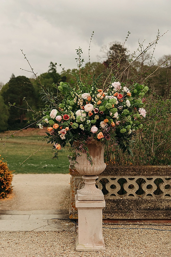 Wedding floral arrangement in a stone urn with blush and peach blooms and arching greenery on a garden pedestal by a gravel path