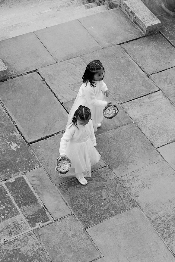 Flower girls in white tulle dresses carry flower girl baskets of petals, walking together across a stone courtyard in fuzzy cardigans