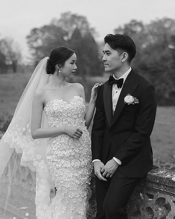Couple portrait in a black and white wedding portrait style, bride in strapless lace gown and veil touching groom on a stone balustrade lawn