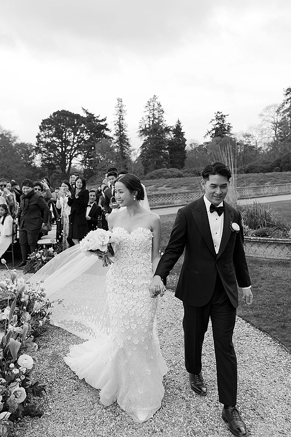 Wedding recessional as bride and groom walk the aisle holding hands, her veil trailing and bouquet in hand on a garden terrace path