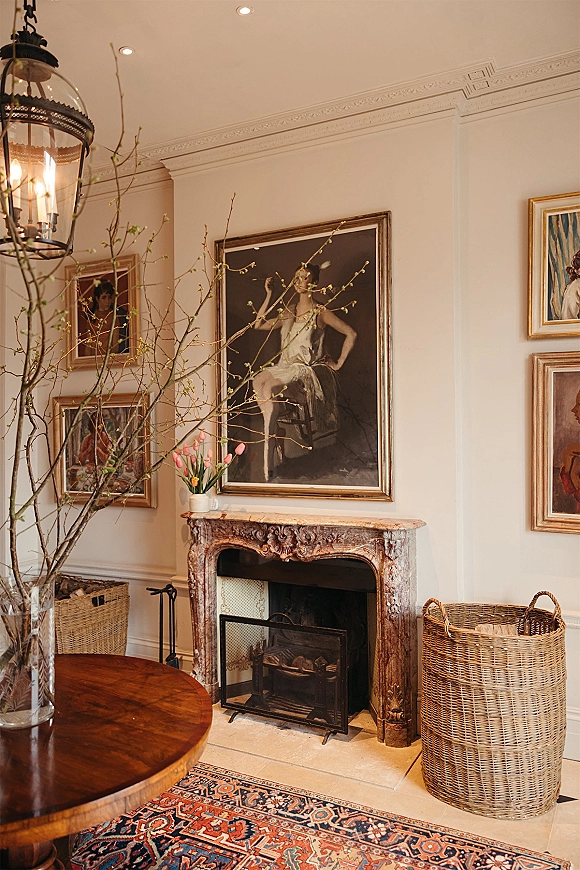 Wedding venue interior with ornate stone fireplace mantel, framed artwork, and a vase of pink tulips on a round wooden table under lantern light