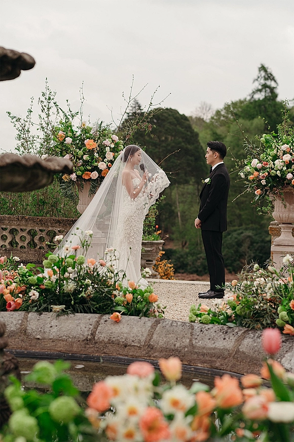 Wedding vows as bride reads to groom, long cathedral veil trailing over lace dress on a stone terrace beside a fountain and gardens