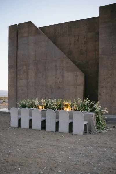 Reception tablescape on an outdoor reception table, lined with candles, set against a concrete wall, desert ground, and an open sky.