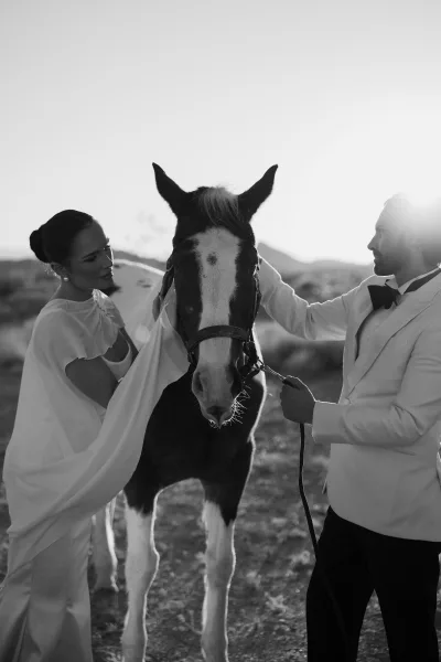 Couple portrait of a bride and groom with horse in a sunlit desert, mountains rising behind them under a wide open sky.